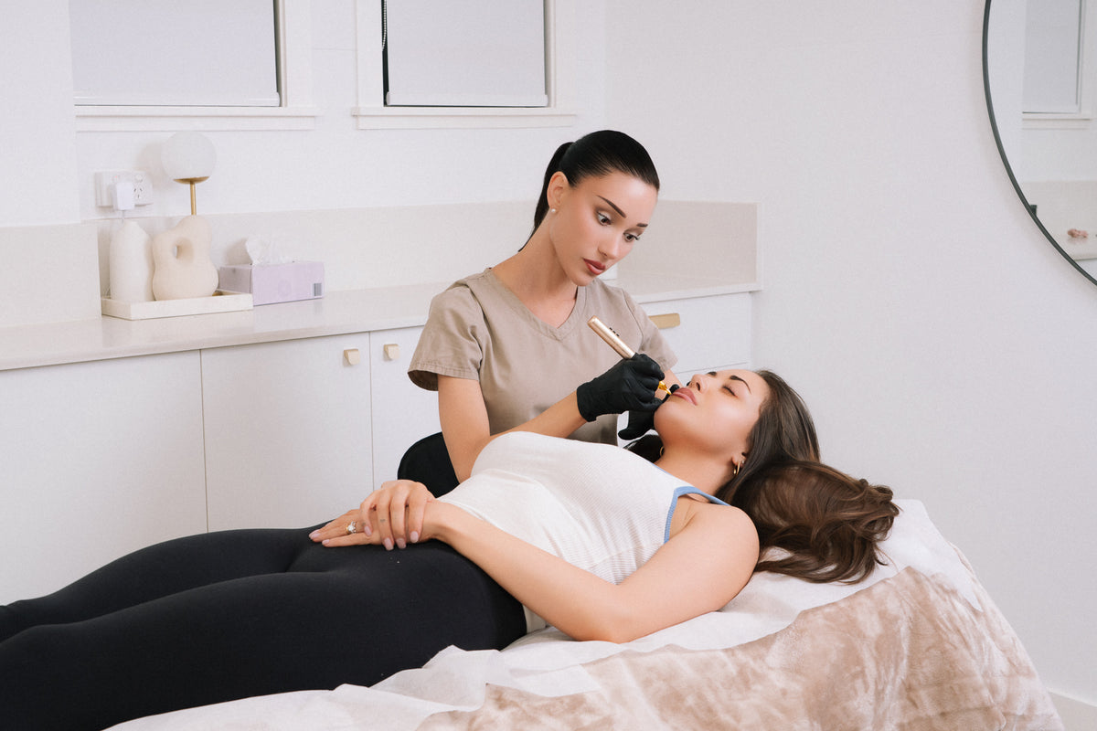A beauty therapist applies cosmetic eyebrow tattoo to a client lying on a treatment bed in a bright, modern spa room.